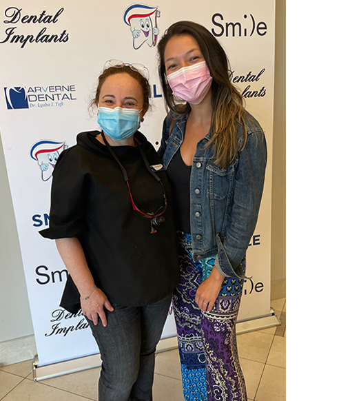 Two women smiling, wearing masks, posing in front of a dental implants banner.