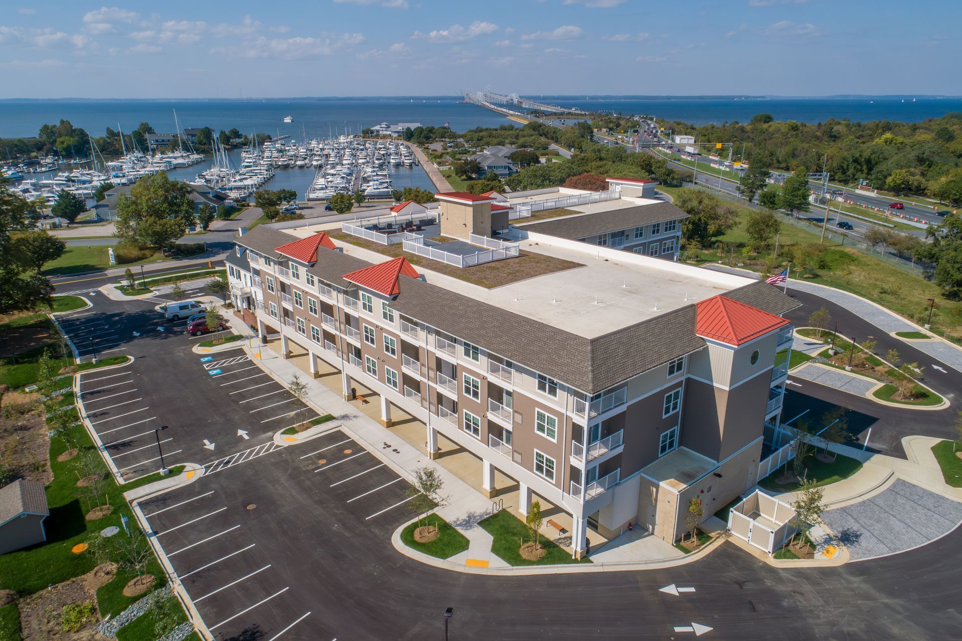An aerial view of a multi-story apartment building with a red-tiled roof, near a marina and a long bridge over water.