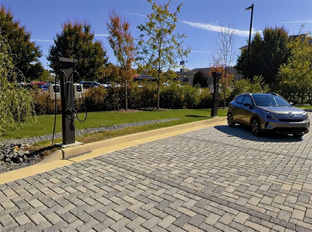 A silver electric vehicle parked in a paved lot next to two EV charging stations on a sunny day.