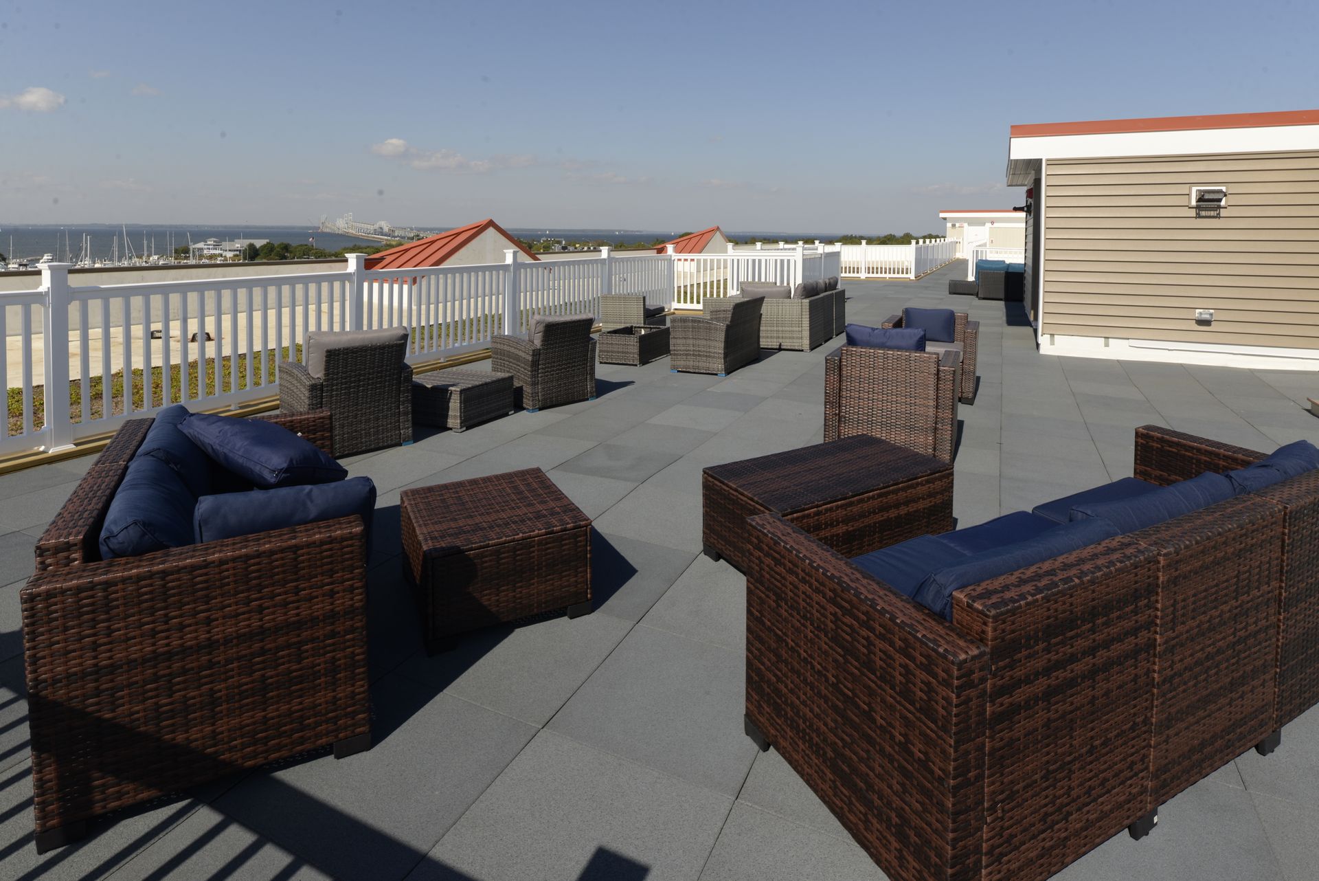 A rooftop patio with wicker furniture and blue cushions, overlooking a beach and the ocean under a clear sky.