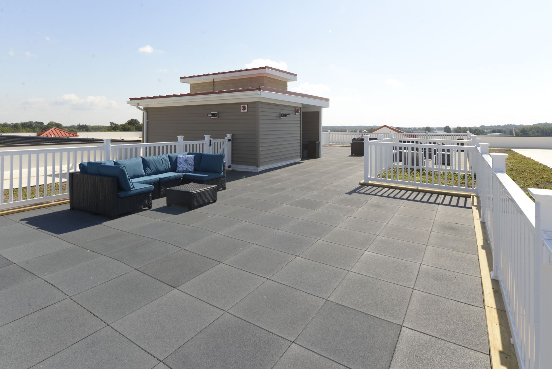 A rooftop patio with grey modular tiles, white railings, a blue sectional sofa, and a small building under a clear sky.