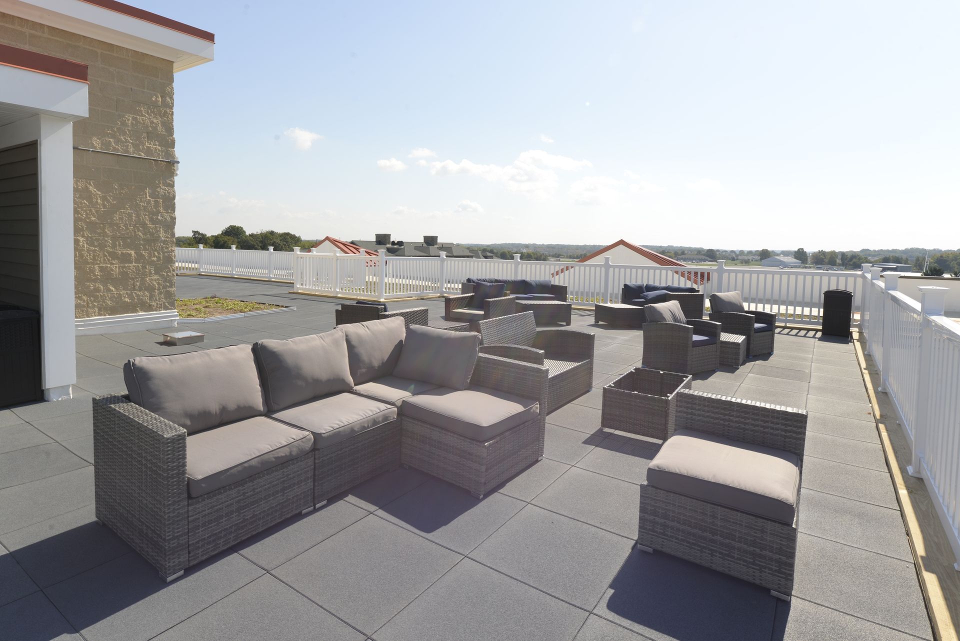 A rooftop patio with gray wicker sectional sofas and chairs on a tiled deck, enclosed by a white fence under a sunny sky.