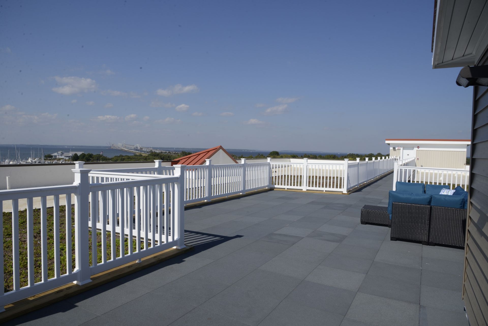 A spacious rooftop patio with grey stone tiles, white railings, and a blue lounge area overlooking the ocean.