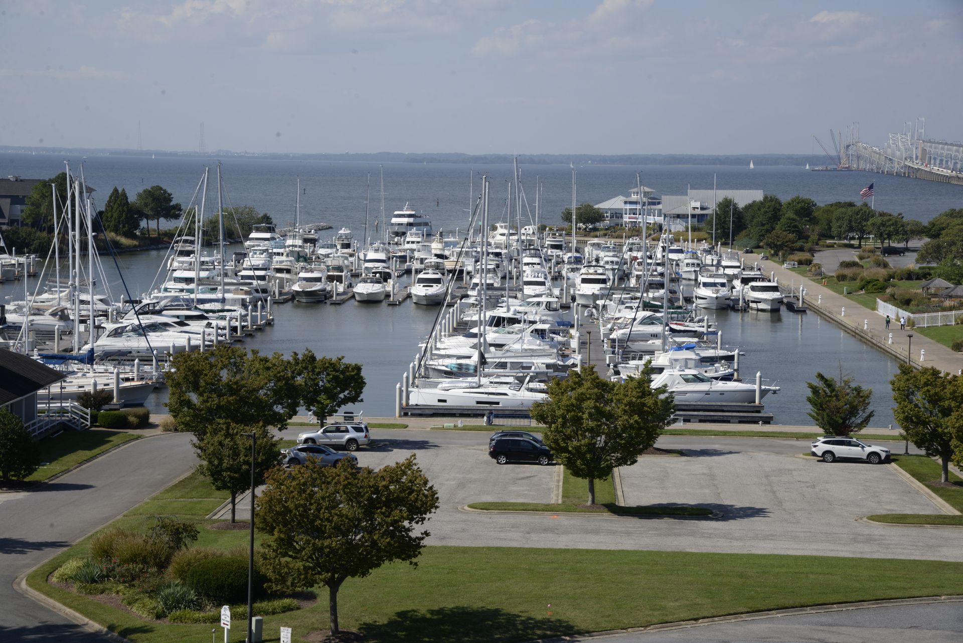 A marina full of docked sailboats and yachts under a clear sky, viewed from an elevated parking lot area.
