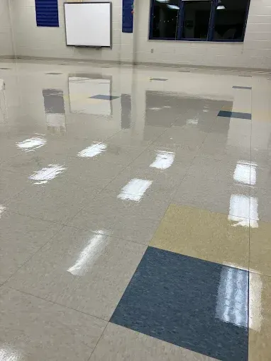 Glossy, tiled floor in a room, with rectangles of lighter and darker colors reflecting light. Whiteboard and windows in the background.
