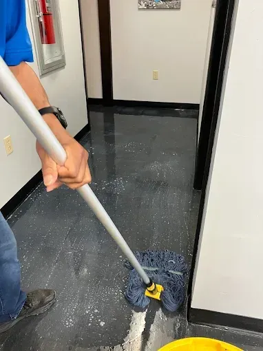 Person mopping a black floor in an office hallway with white walls and black trim.