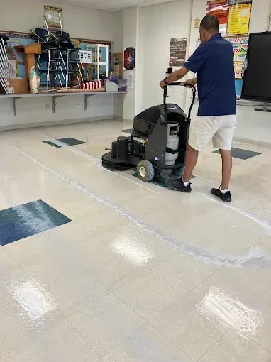 Person operating a floor cleaner in a room with light-colored flooring and blue tile accents.