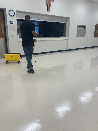 A person mopping a shiny, white floor in a brightly lit room. A yellow bucket on wheels is nearby.