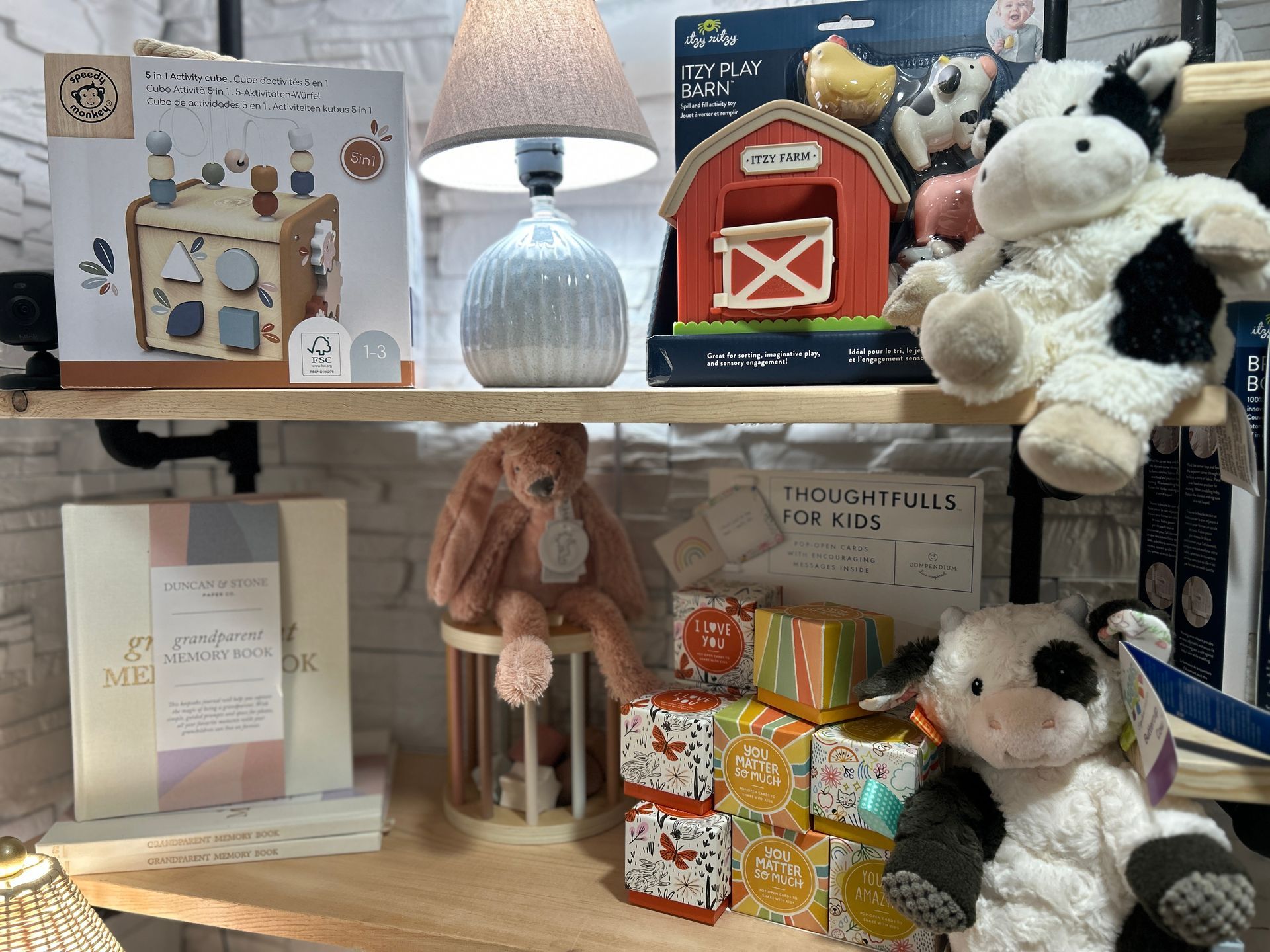 Shelf display with a lamp, plush cows, greeting cards, and colorful gift boxes in a cozy shop setting
