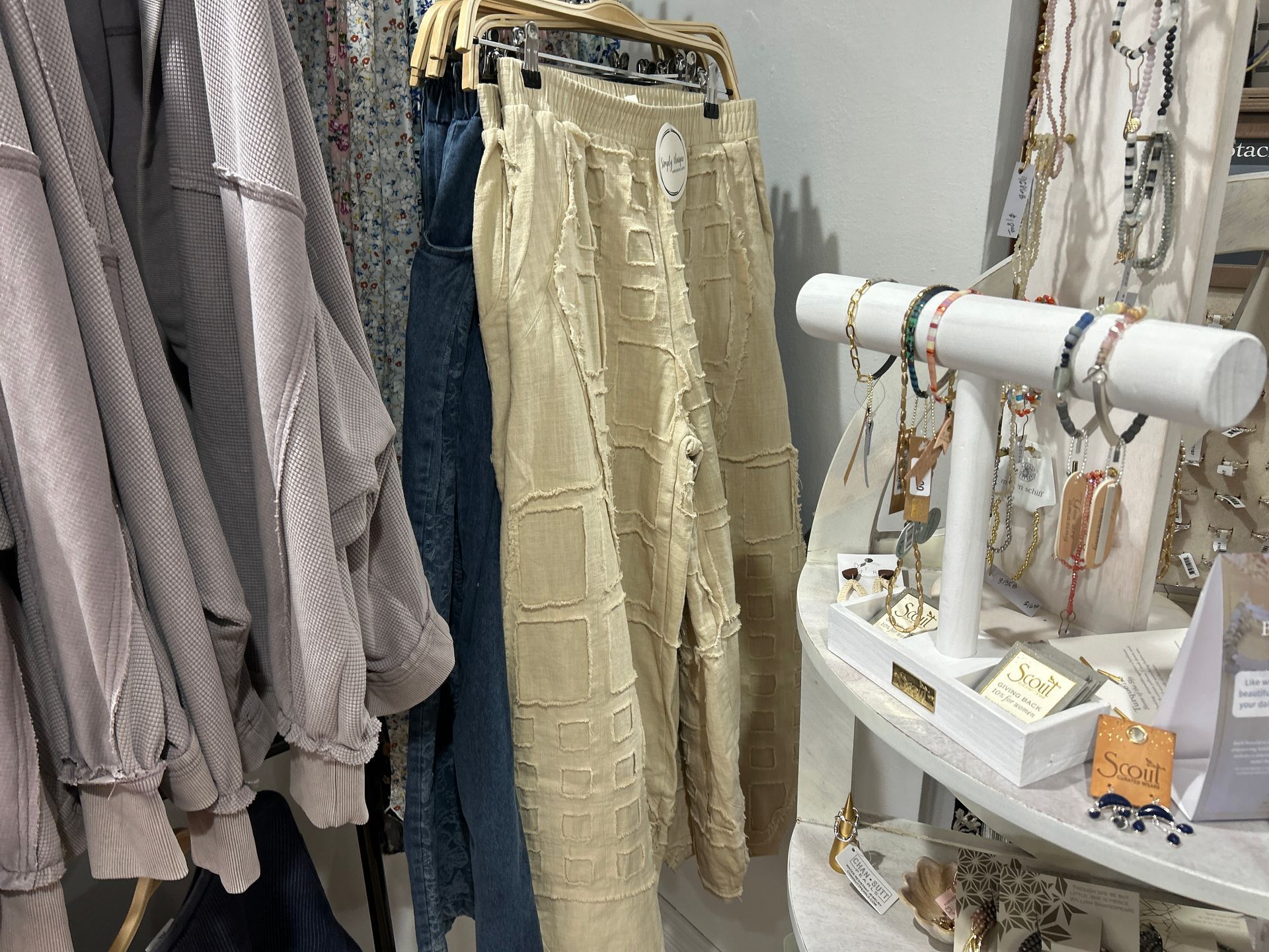 Clothing rack with beige pants and folded tops beside a white jewelry display stand in a boutique