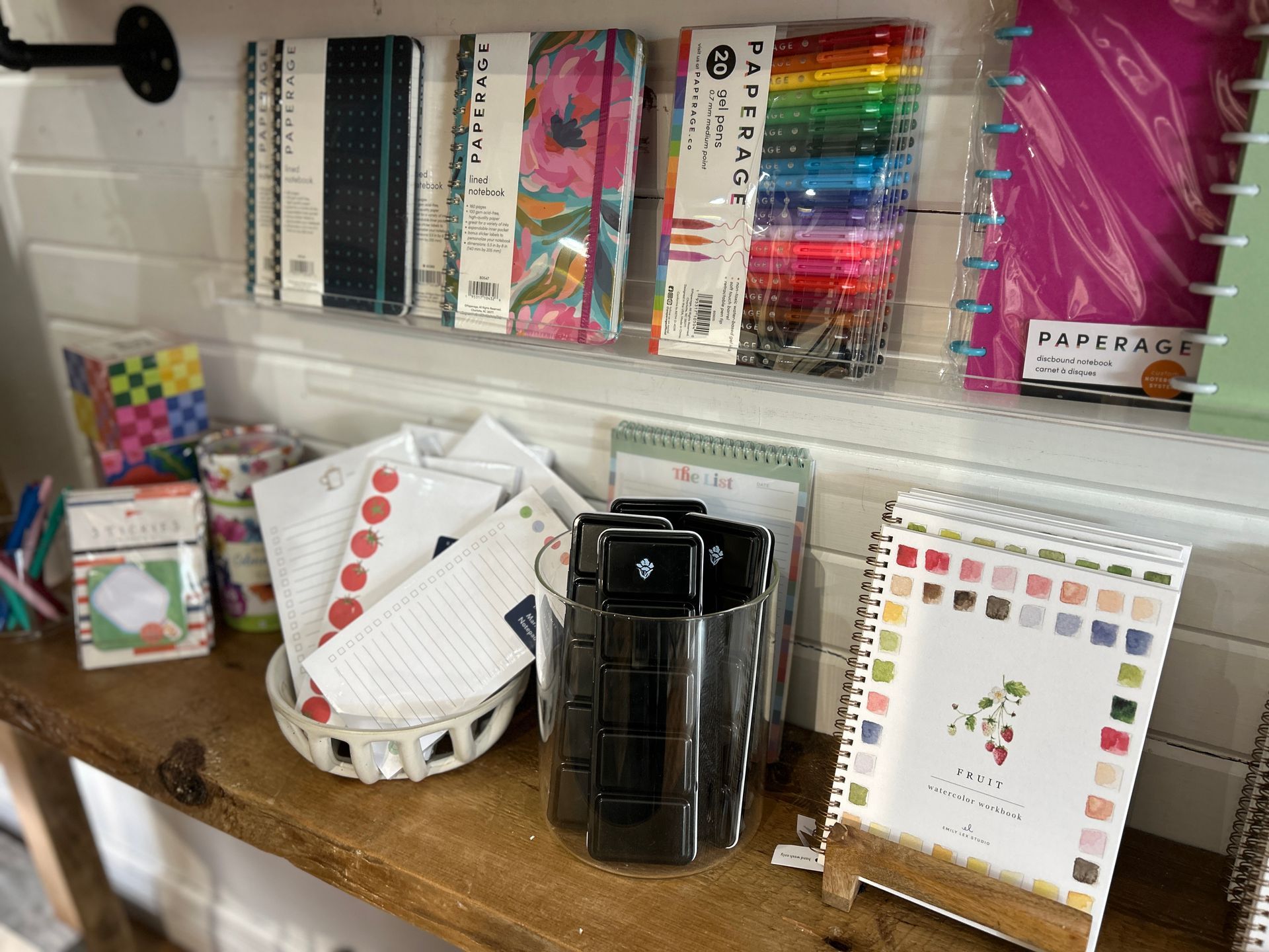 Craft supplies displayed on a shelf, including pens, notebooks, sticky notes, and a black mug or tumbler.