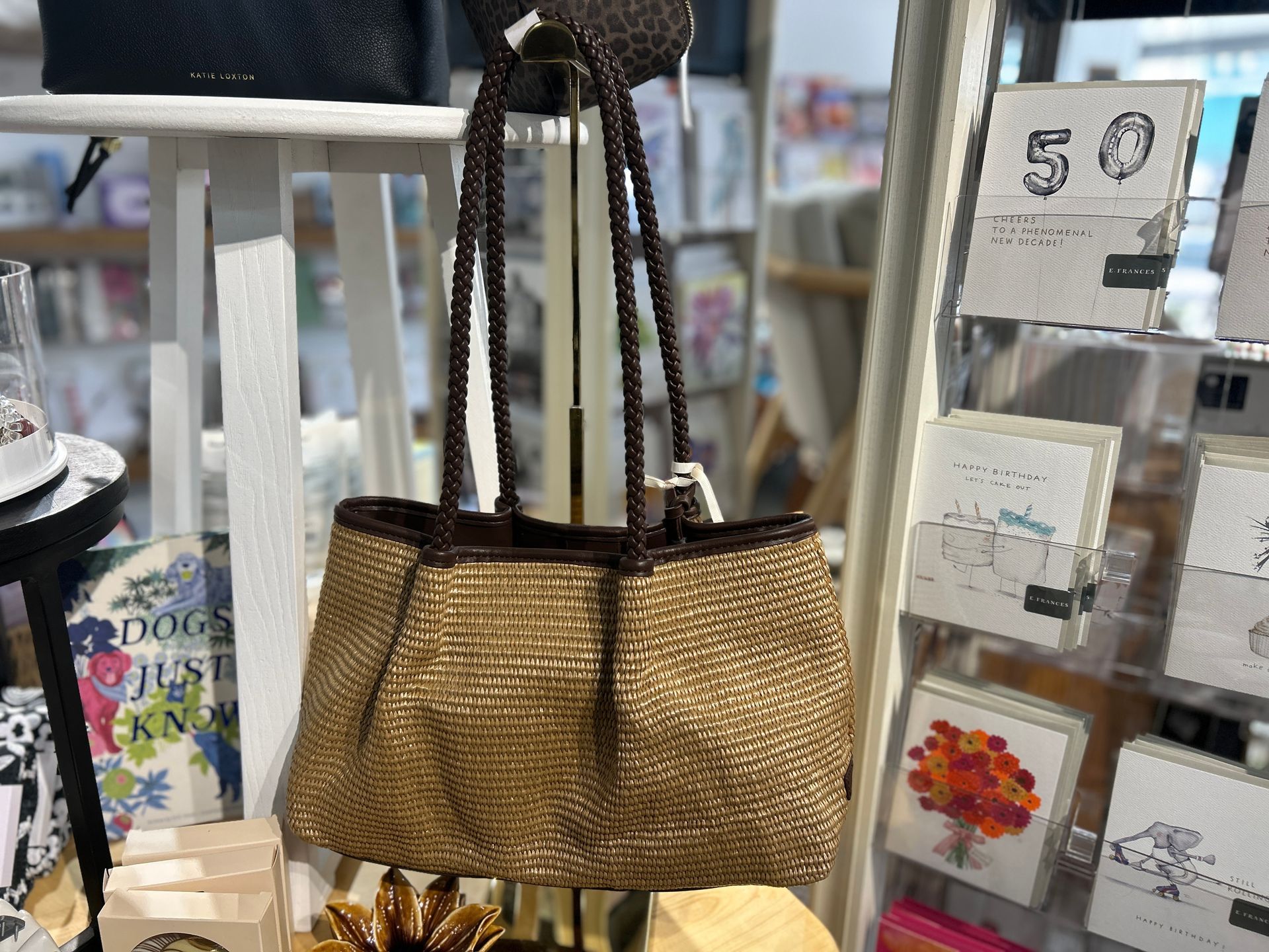 Straw tote bag with black handles hanging in a store display, with greeting cards and price tags nearby