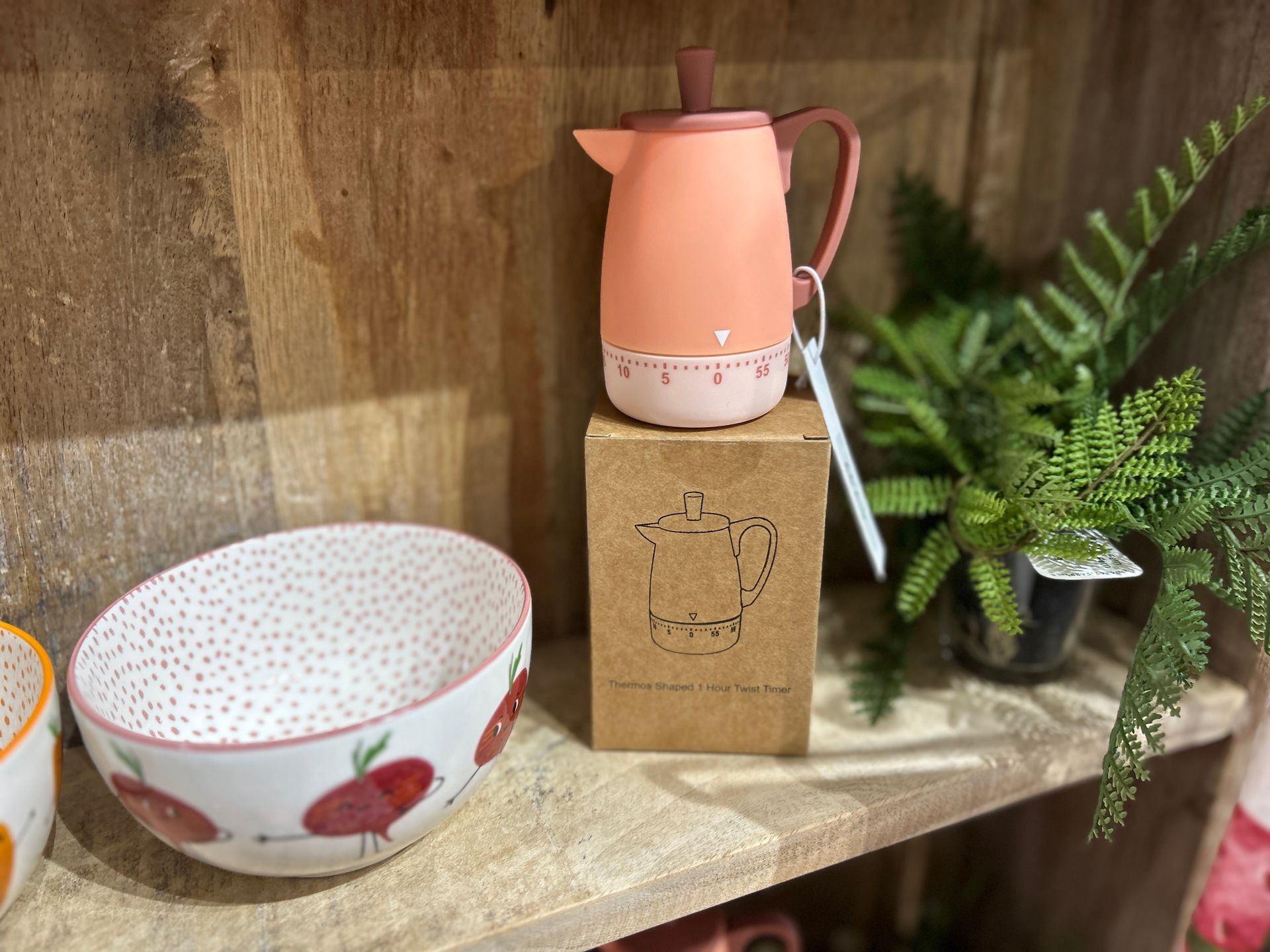 Pink pitcher on a kraft box beside patterned bowls and a fern on a wooden shelf