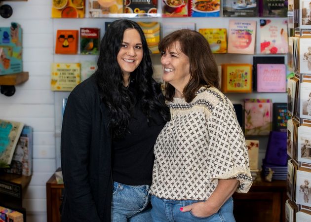 Two women smiling in front of a colorful card display in a shop