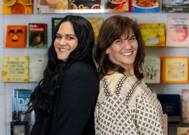 Two women smiling back-to-back in front of a colorful wall of framed artwork and prints