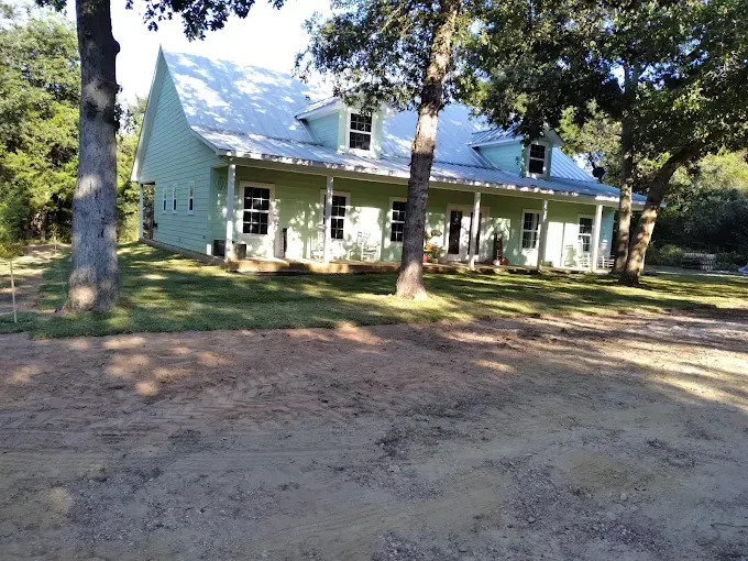 Light blue farmhouse with white porch, surrounded by trees and dirt yard in daylight