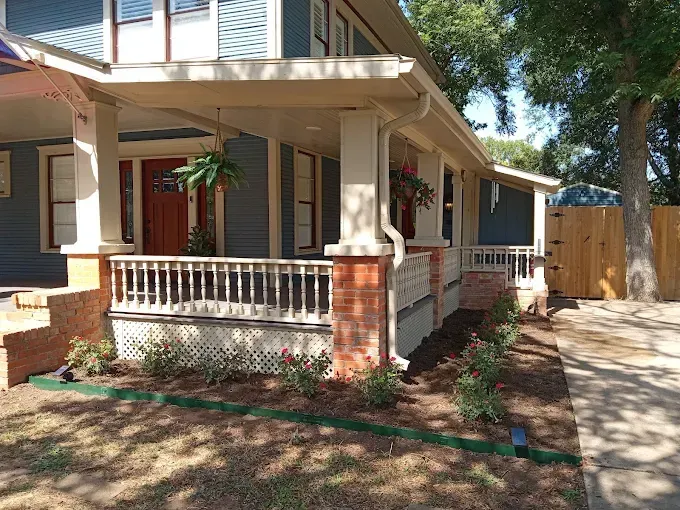 Front porch of a house with brick columns, white railing, and a shaded walkway lined with trees