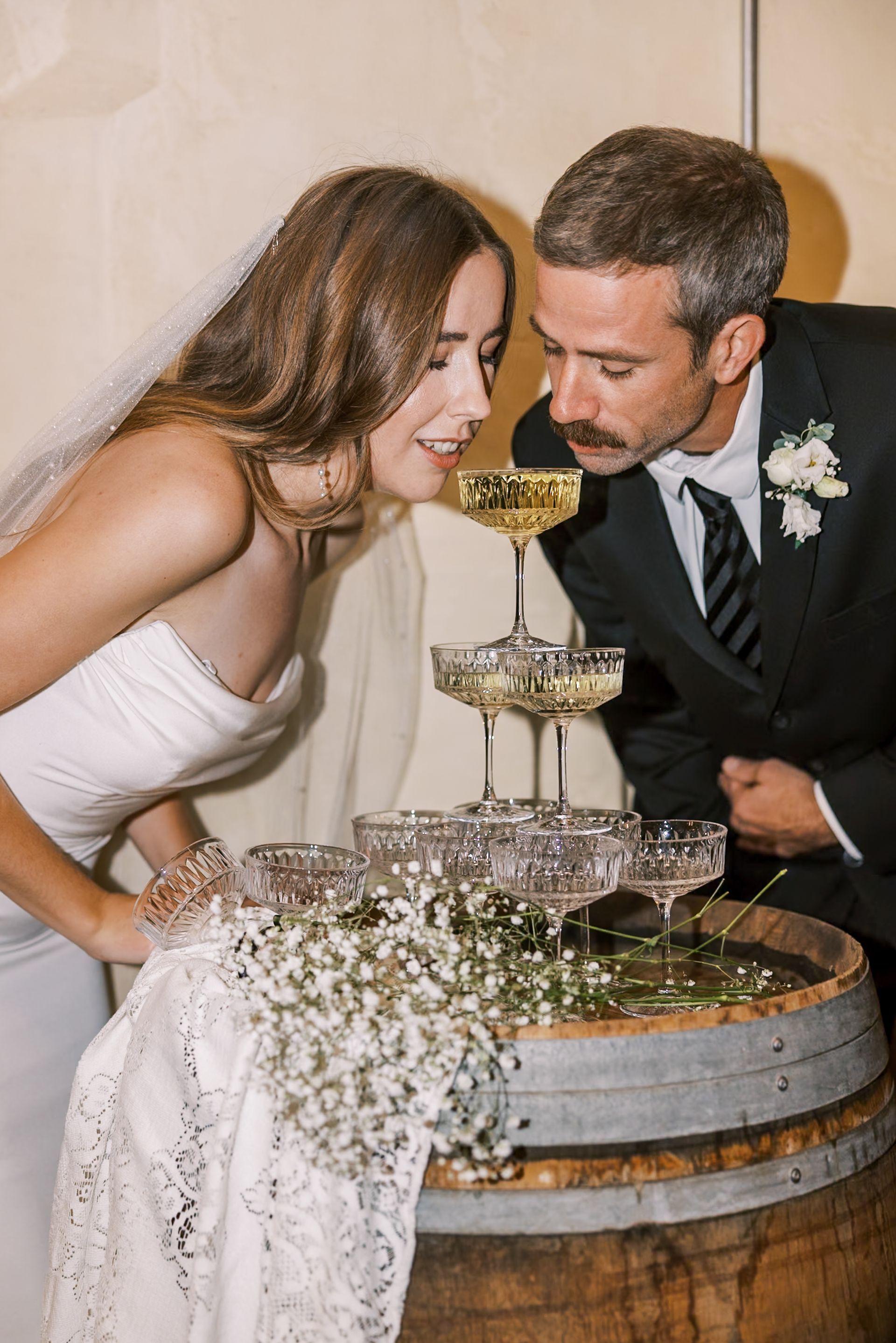 Bride and groom leaning toward a champagne tower beside a decorated barrel at a wedding reception