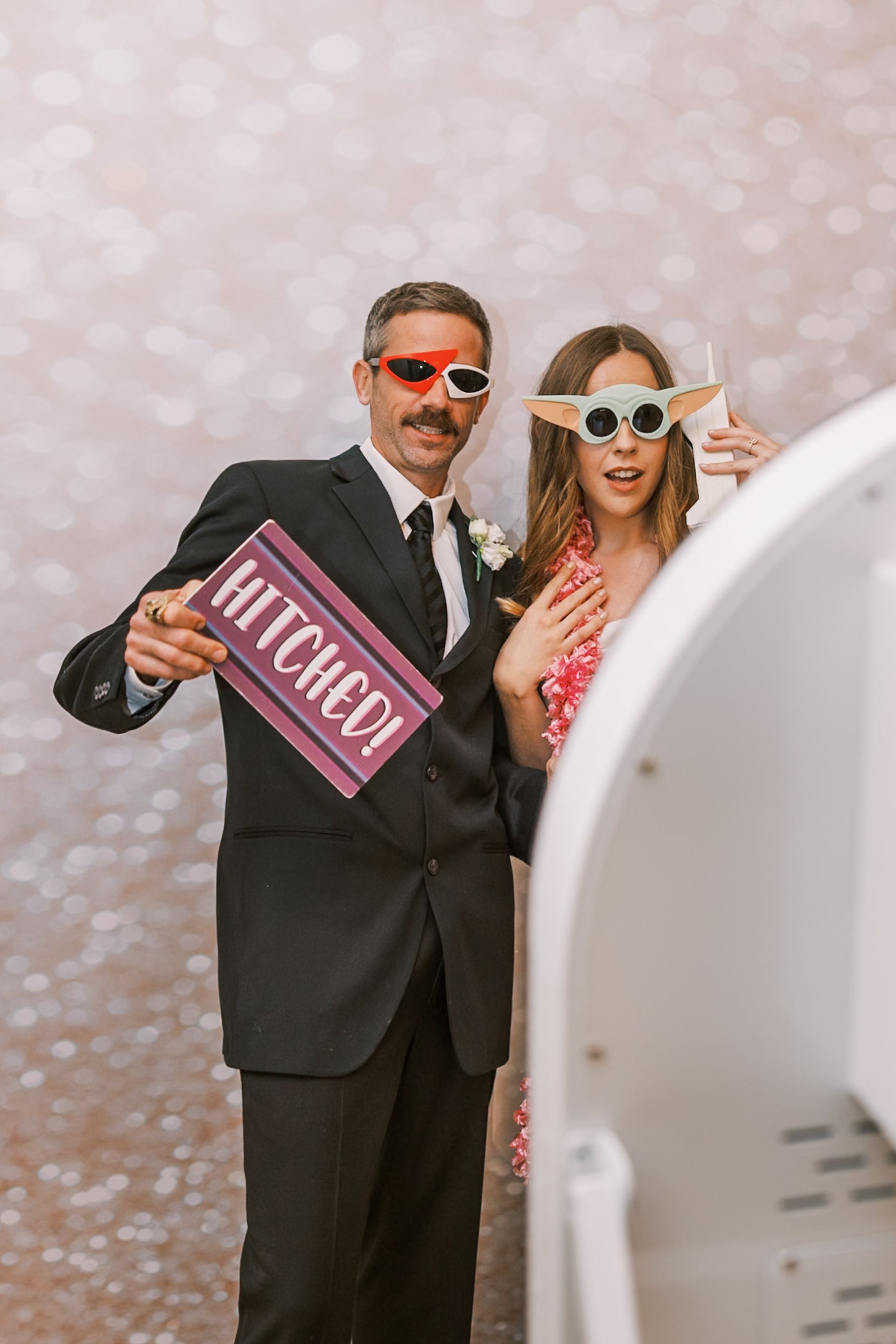 Wedding couple posing with sunglasses and a pink “MARRIED” sign against a blush backdrop