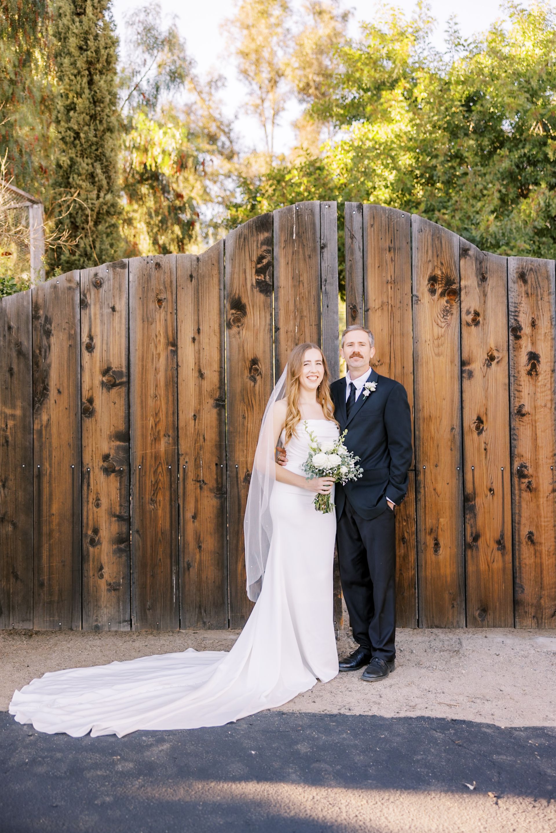 Bride and groom in white and black formalwear posing before a wooden fence outdoors