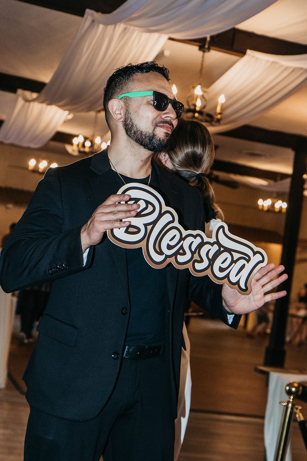 Man in a black suit wearing neon sunglasses and holding a “Blessed” sign at an indoor event
