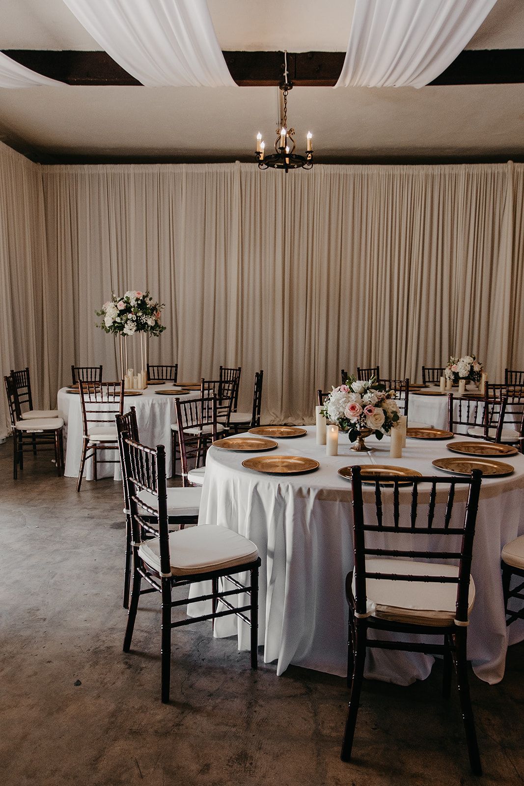 Round tables and black chairs in a decorated banquet hall with white drapes and floral centerpieces