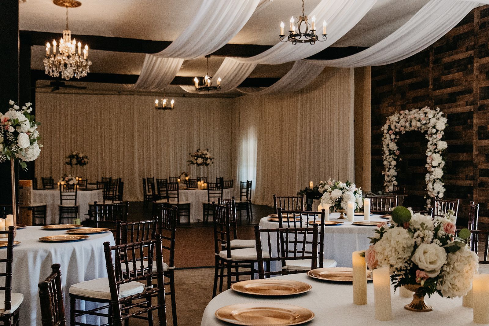 Elegant banquet hall with round tables, chandeliers, draped ceiling fabric, and floral centerpieces
