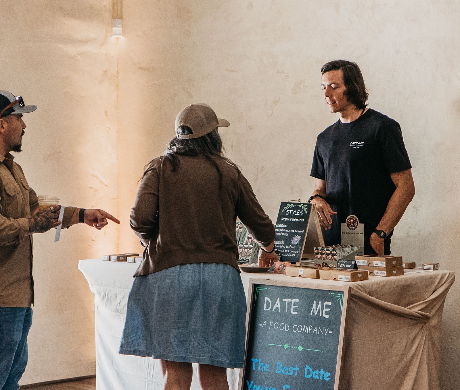 Three people talking at a vendor table with a chalkboard sign reading “DATE ME” and date trays.