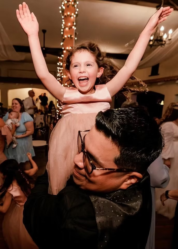 Smiling child with arms raised, lifted onto an adult’s shoulders at a lively indoor celebration