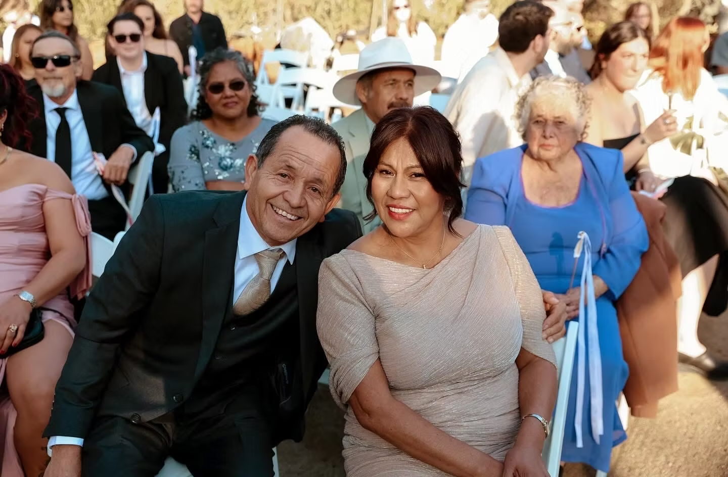 Smiling couple seated outdoors at a formal event, with guests in the background