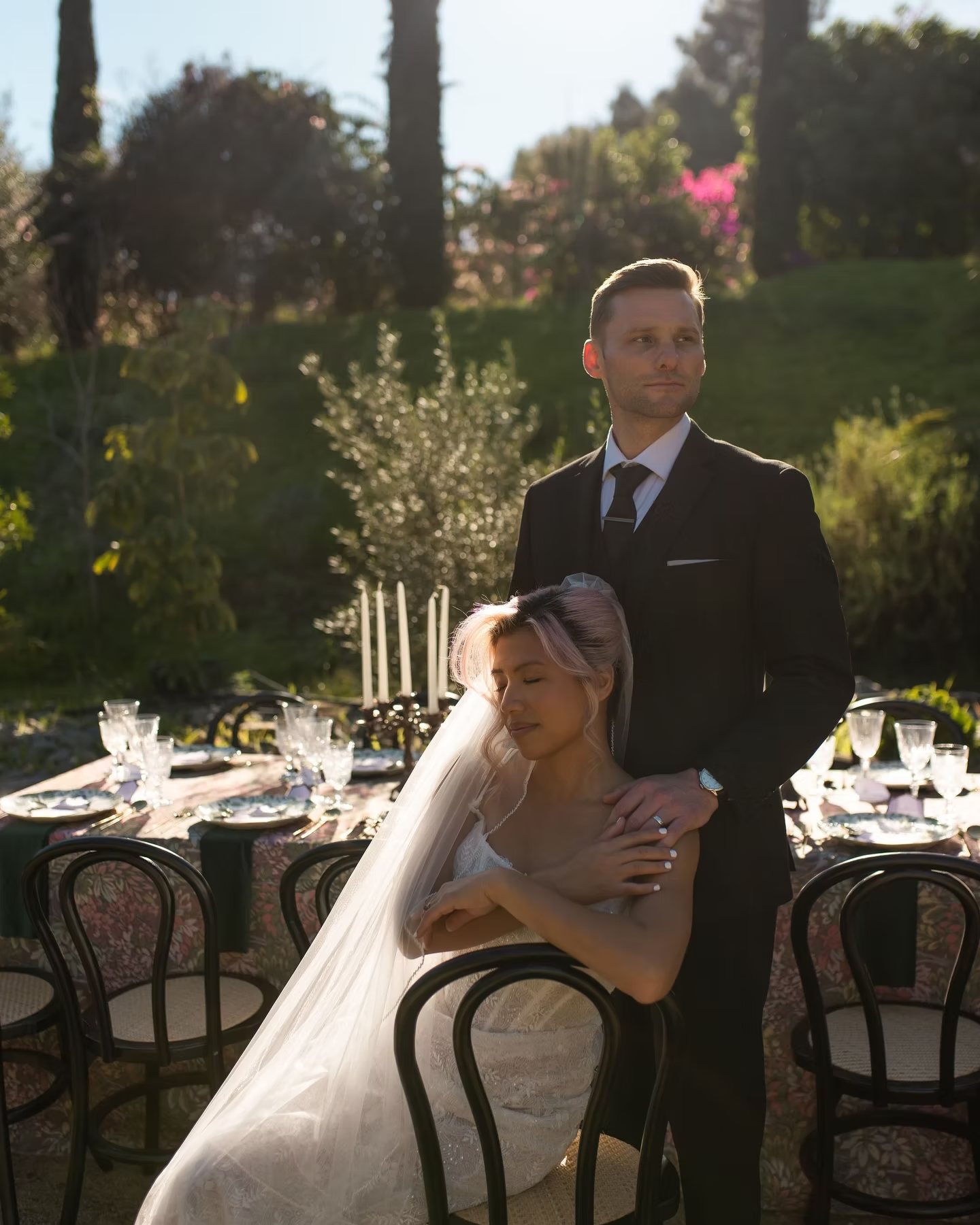 Bride in a white veil sits with groom in a black suit at an outdoor reception table in sunlight