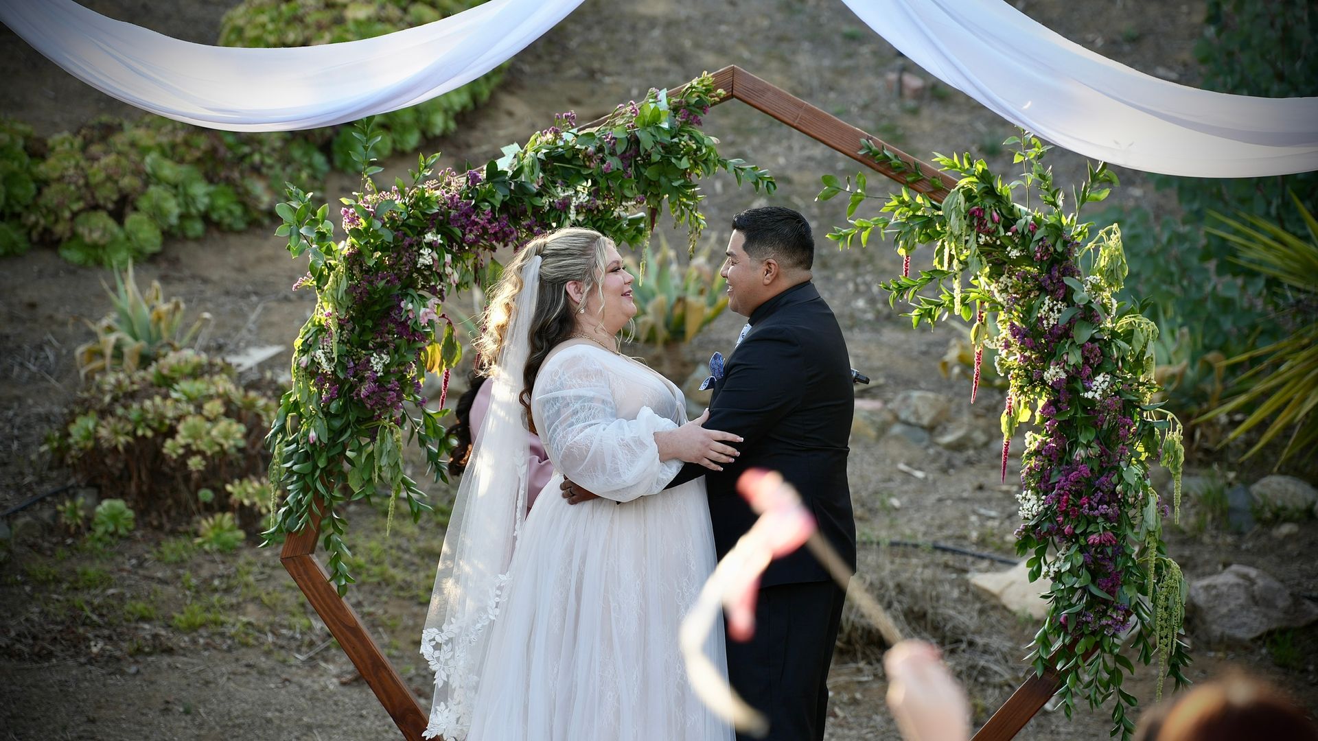 Bride and groom holding hands beneath a floral hexagonal arch at an outdoor wedding ceremony