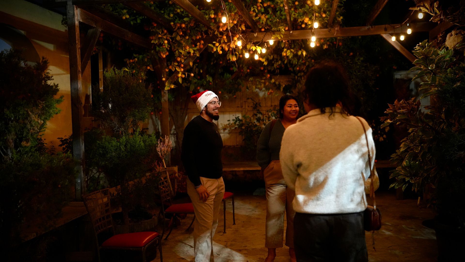 Three people stand in a warmly lit garden patio under string lights at night.