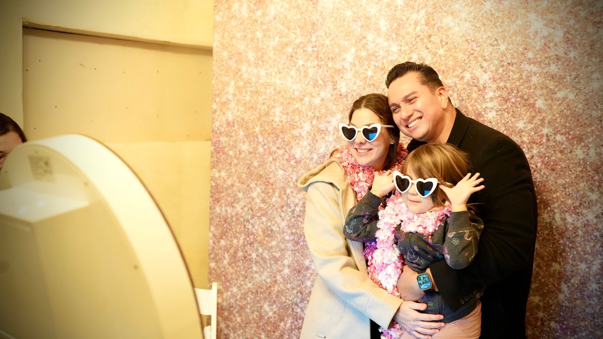 Three people posing in sunglasses for a photo booth against a glittery pink backdrop