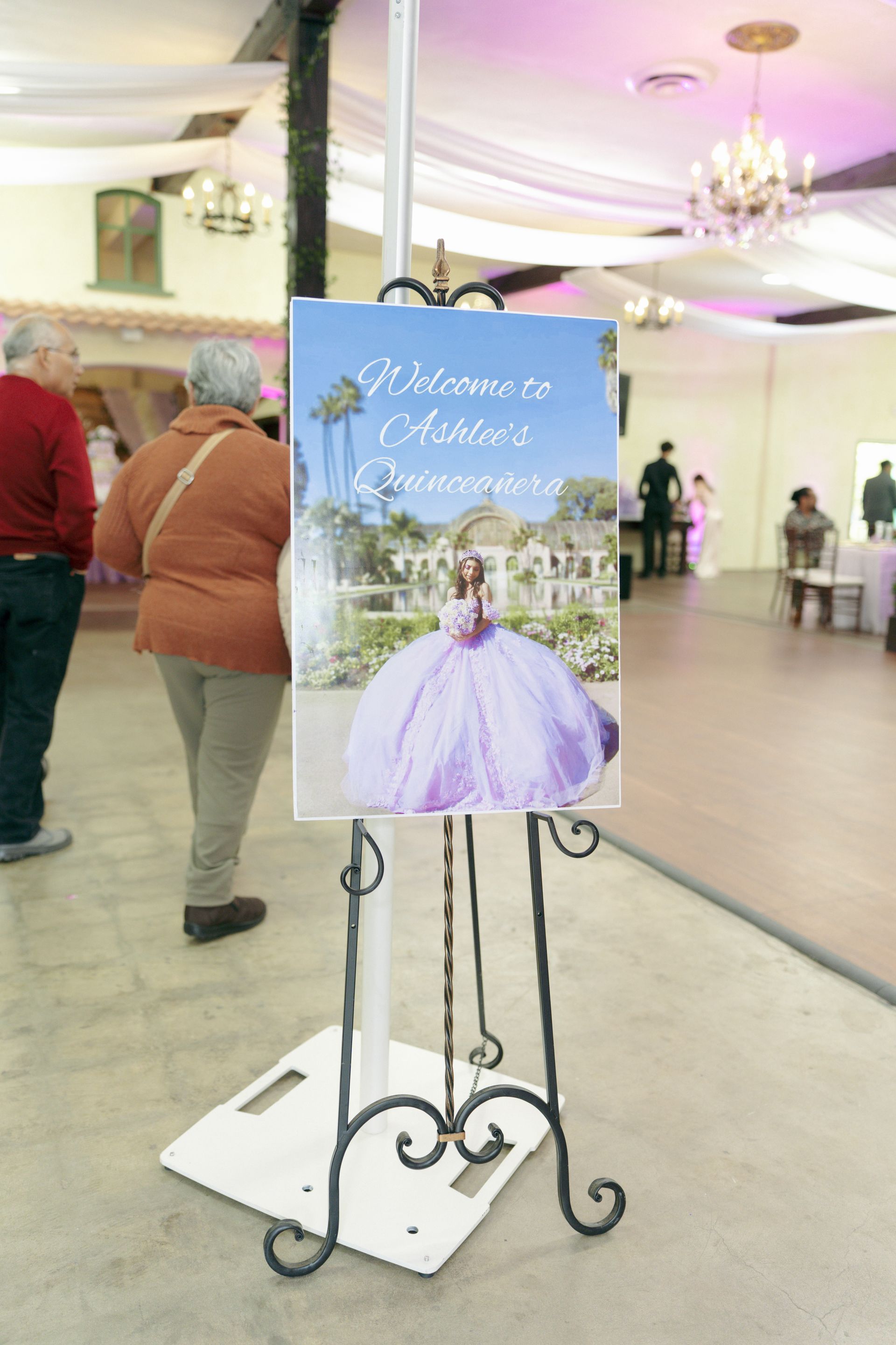 Quinceañera welcome sign on an easel in a decorated hall with guests and purple lighting