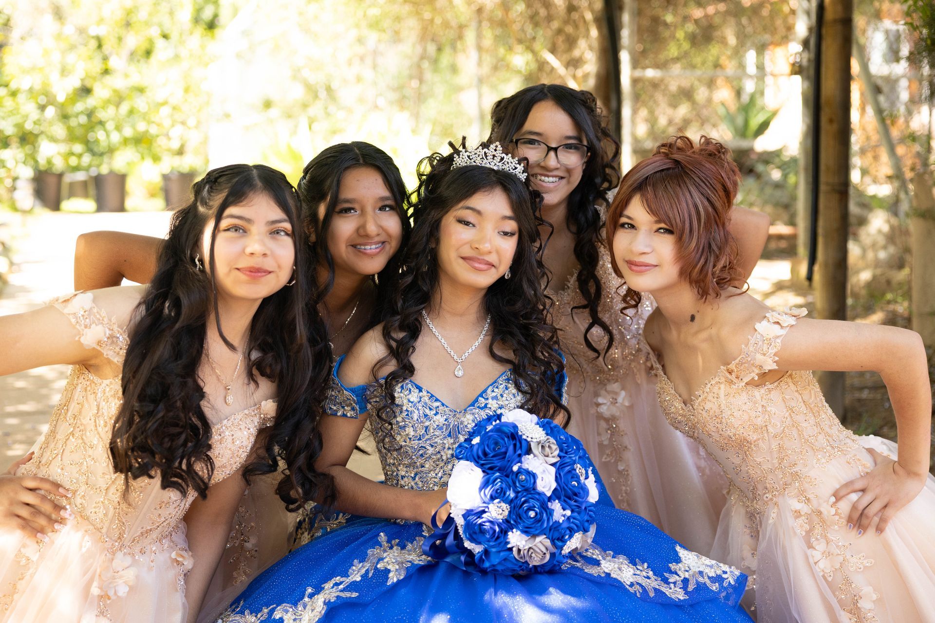 Five women in formal dresses pose outdoors, with one holding a blue-and-white bouquet.