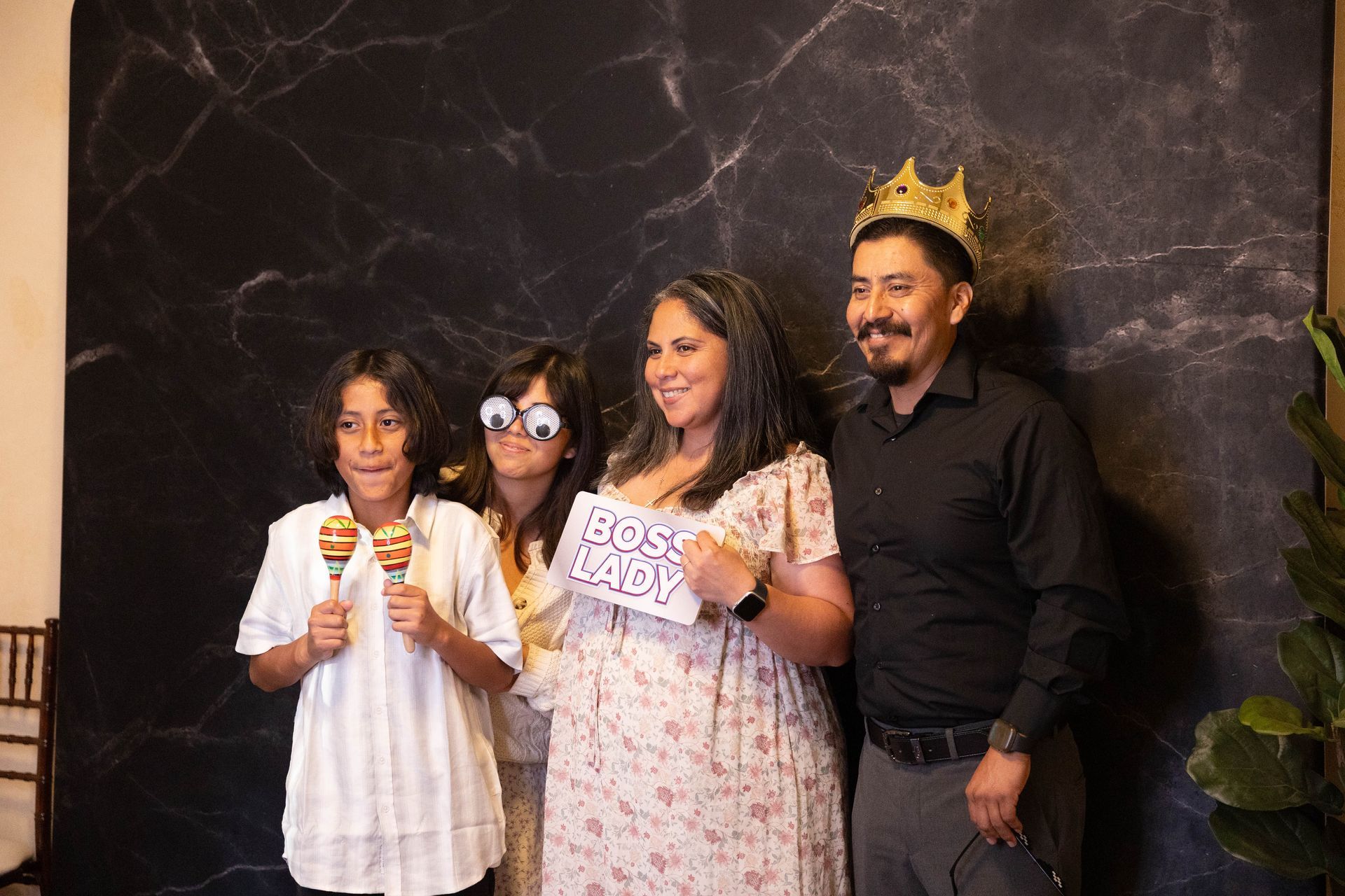 Four people posing indoors in front of a dark marble wall, one wearing a gold crown and all smiling.