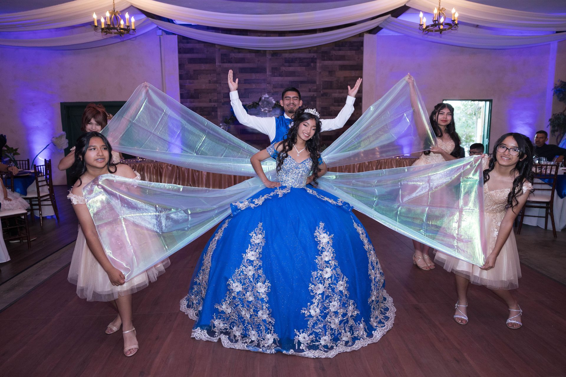 Five dancers in blue and white costumes pose on a decorated stage with draped fabric and purple lighting