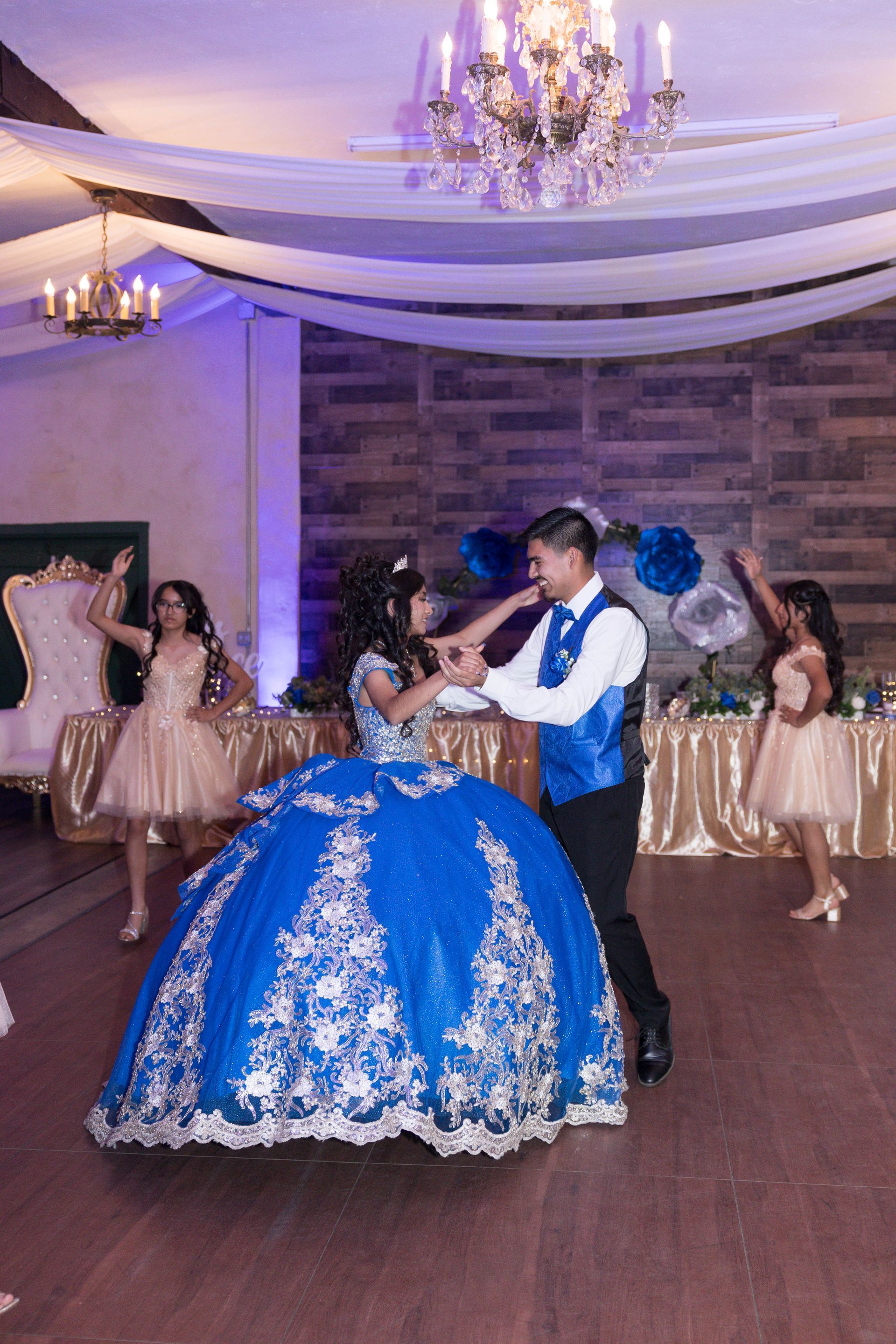 Couple dancing in a decorated ballroom, the woman in a blue gown and the man in a blue vest.