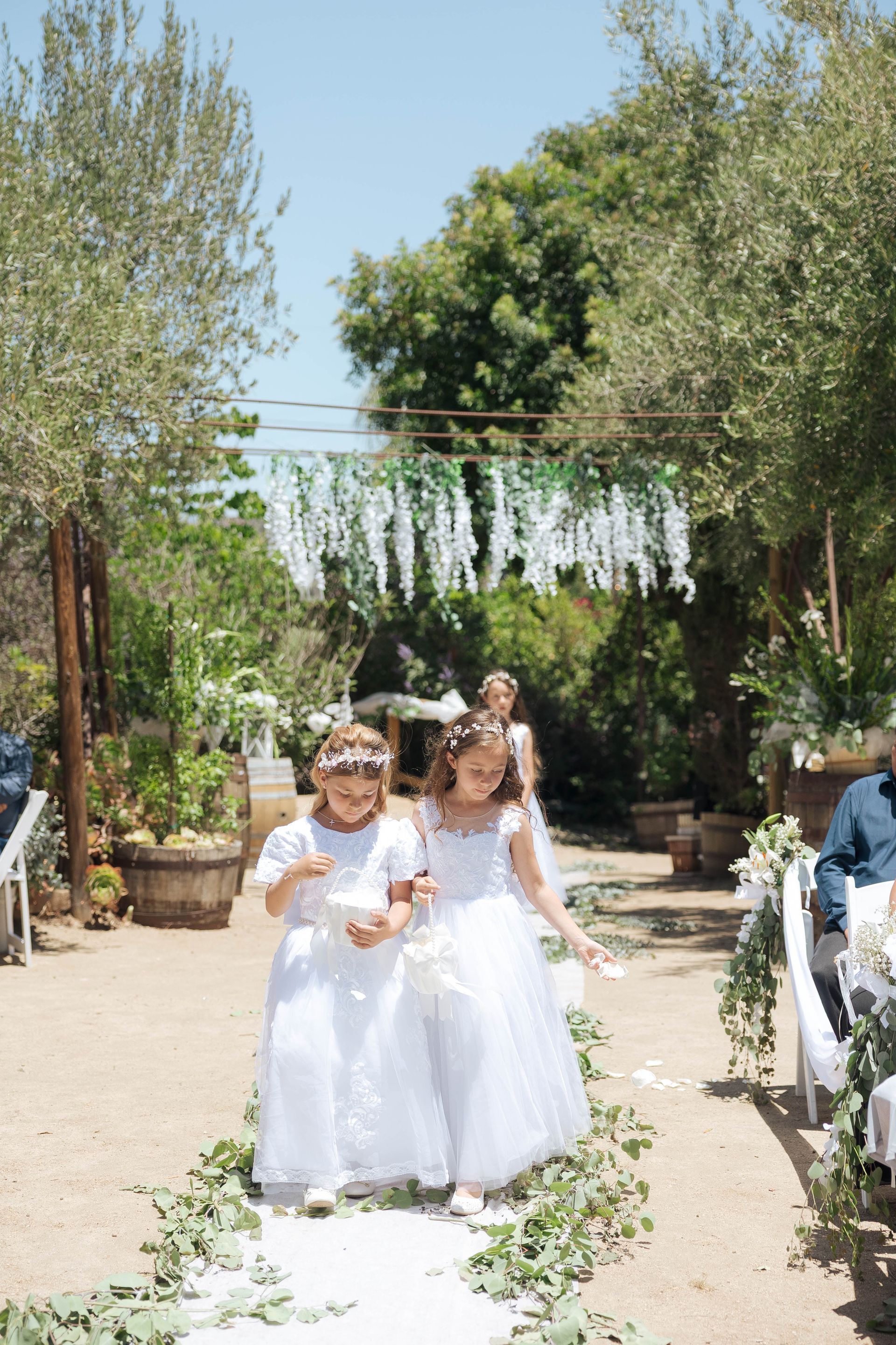 Two children in white dresses walk down a flower-strewn garden aisle under greenery and string lights.