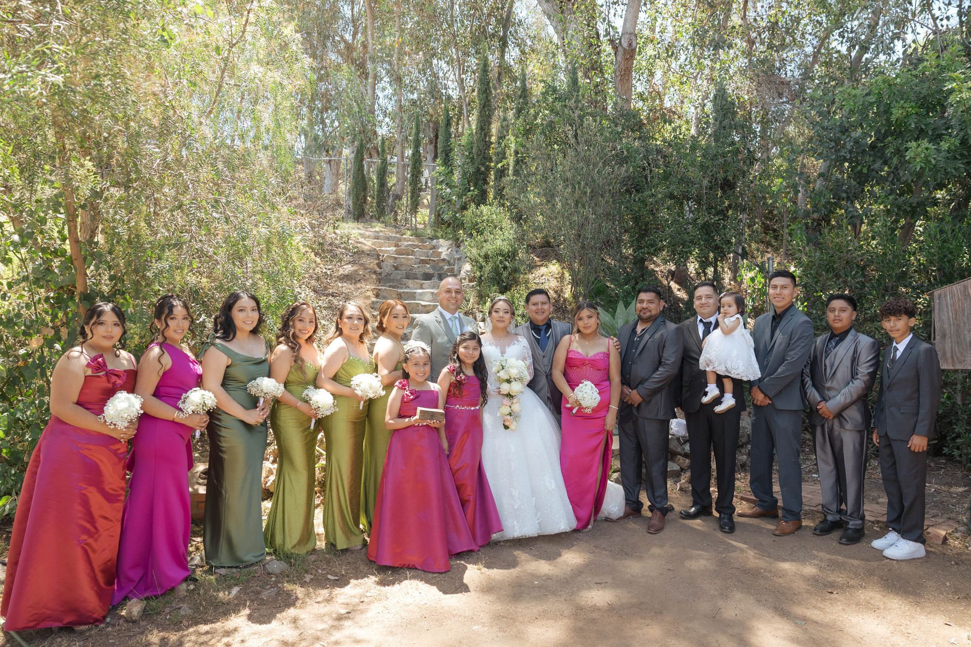 Wedding party posing outdoors in formal attire, with bridesmaids in pink and gold and groomsmen in gray suits.