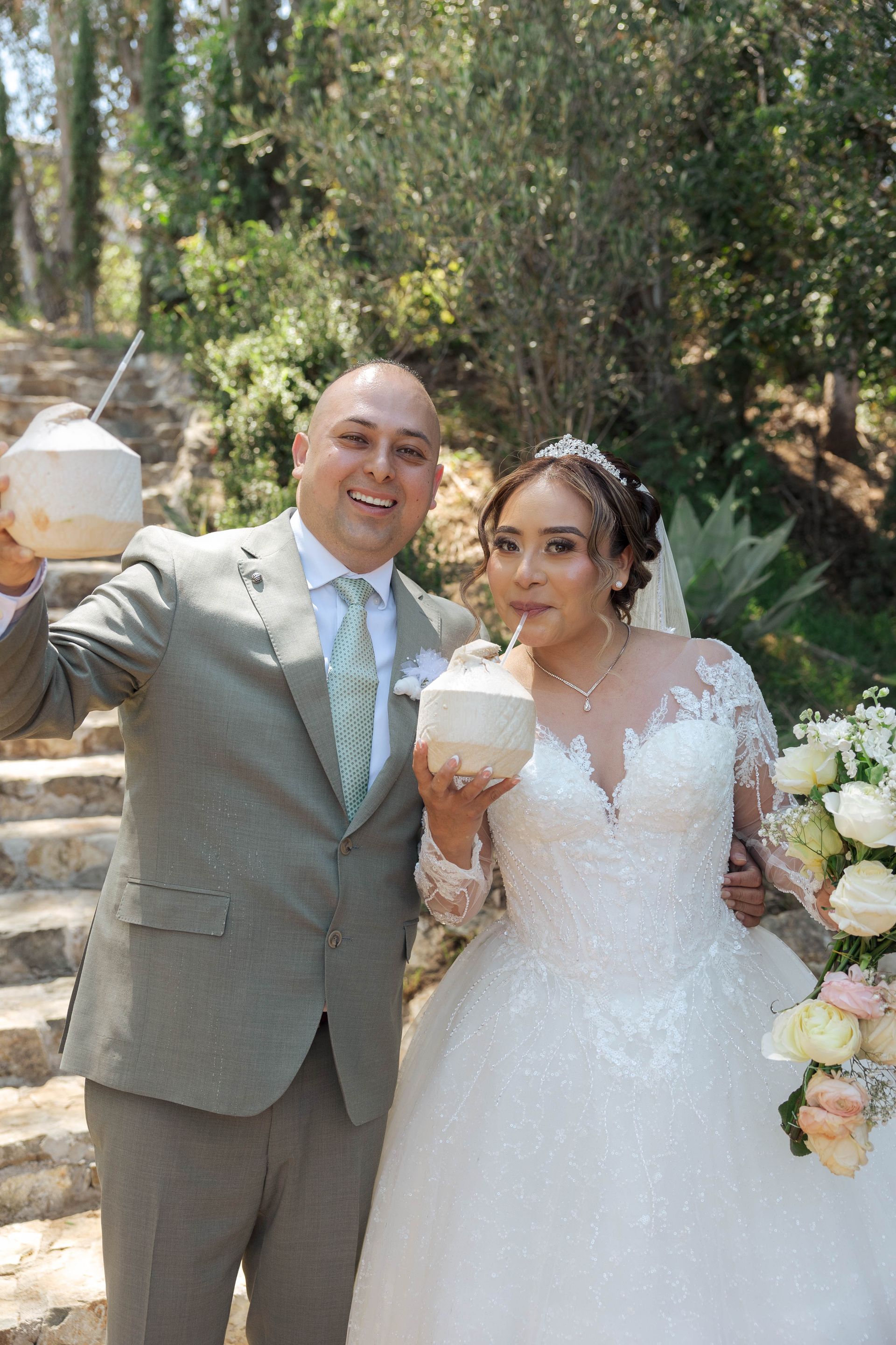 Newlyweds holding coconut drinks in a garden wedding, smiling beside a floral bouquet and greenery.