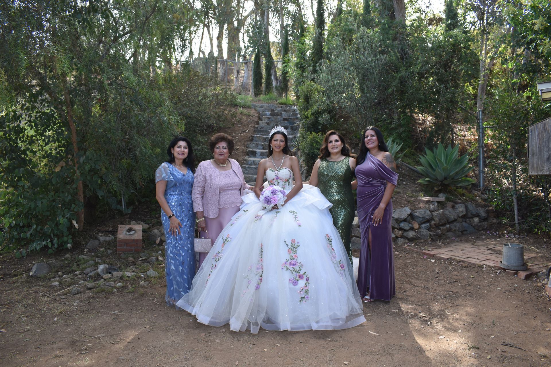 Bride in a white gown with four women in colorful dresses outdoors among trees
