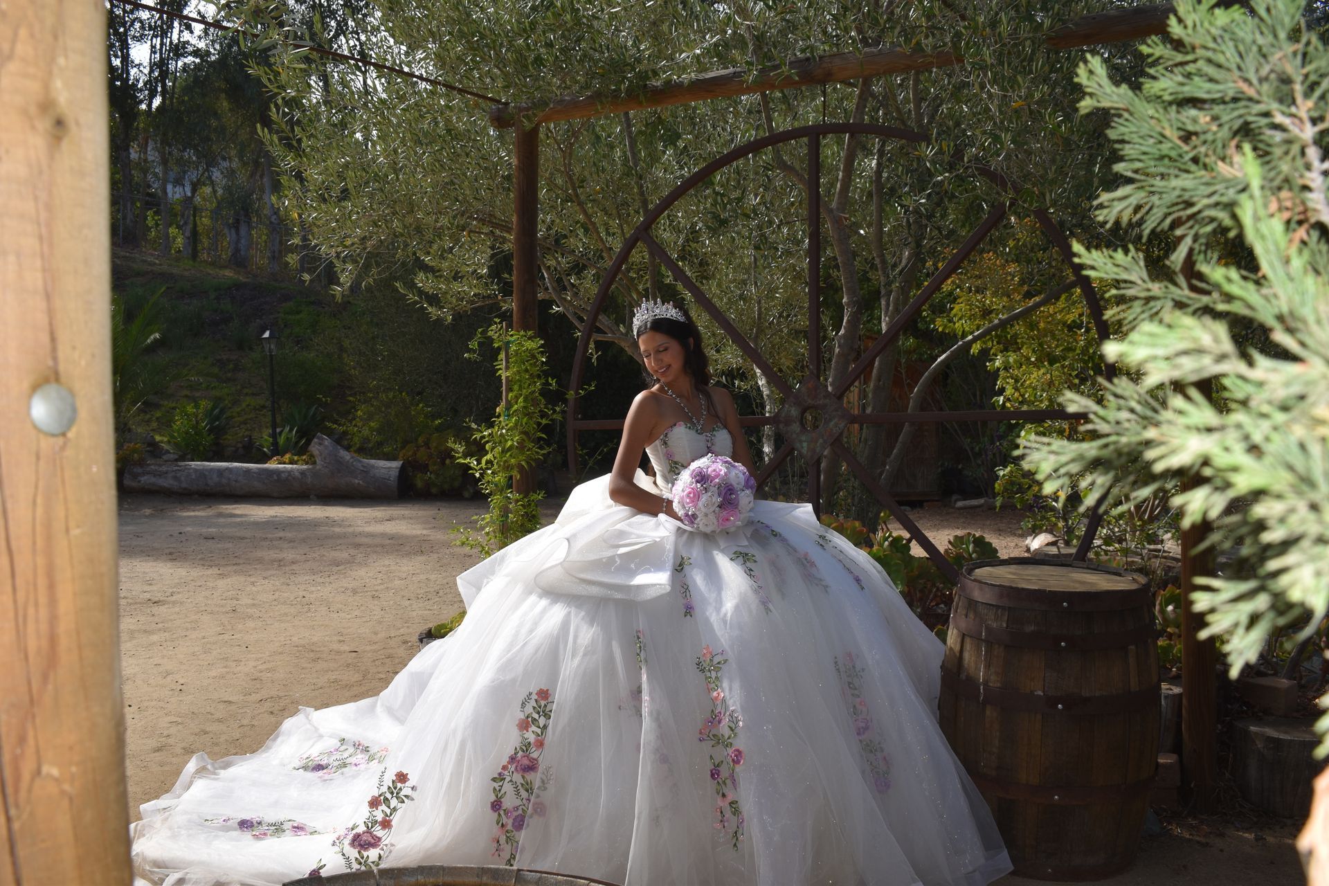 Bride in a white gown holding a bouquet, seated outdoors under a wooden archway.