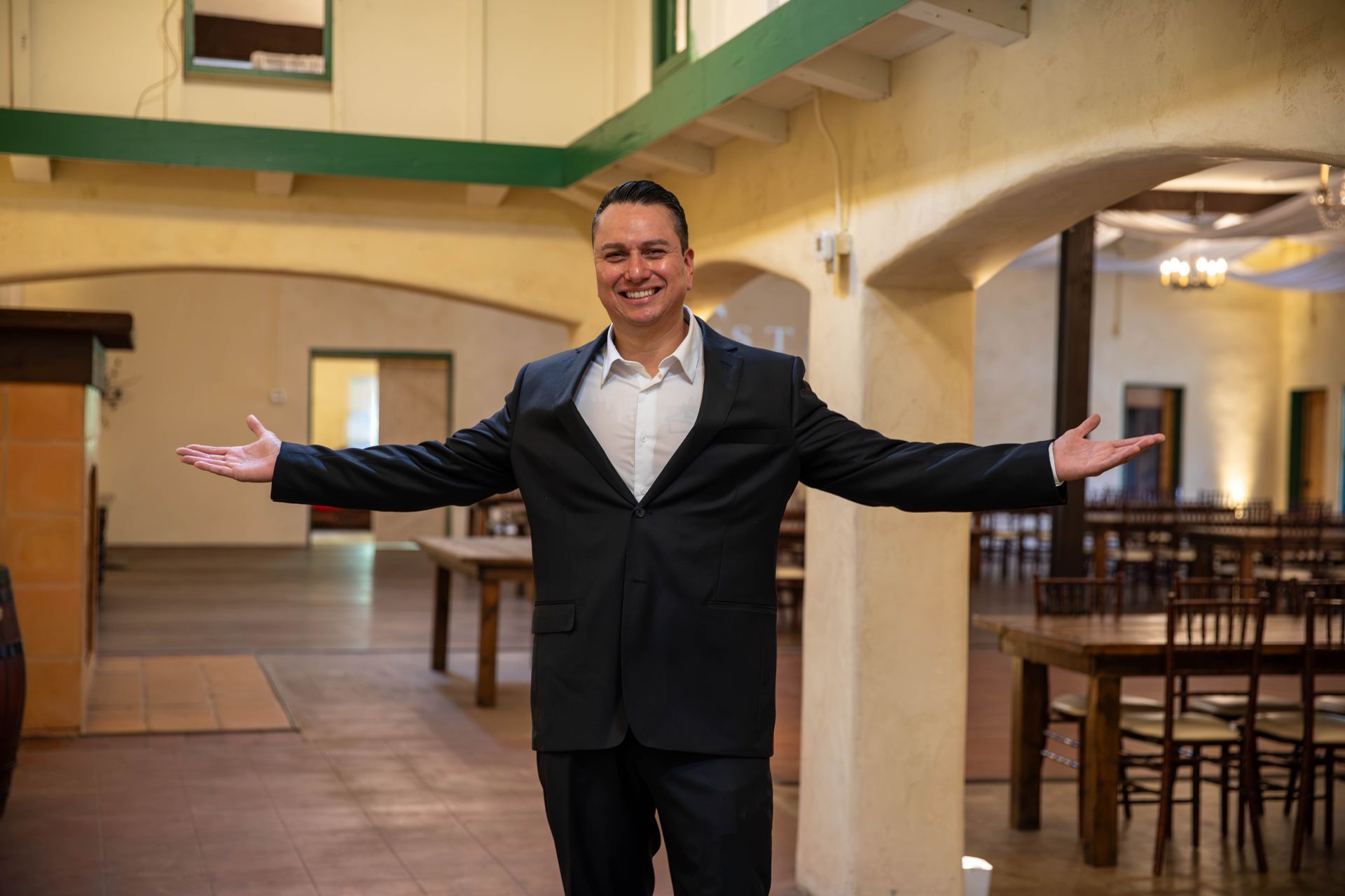 Man in a black suit smiling with arms outstretched in a spacious indoor hall