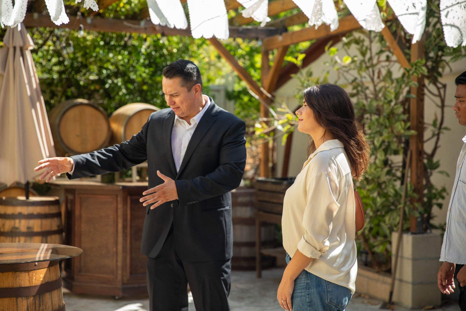 Two people talking in a sunny outdoor patio with wooden barrels and barrels stacked behind them.