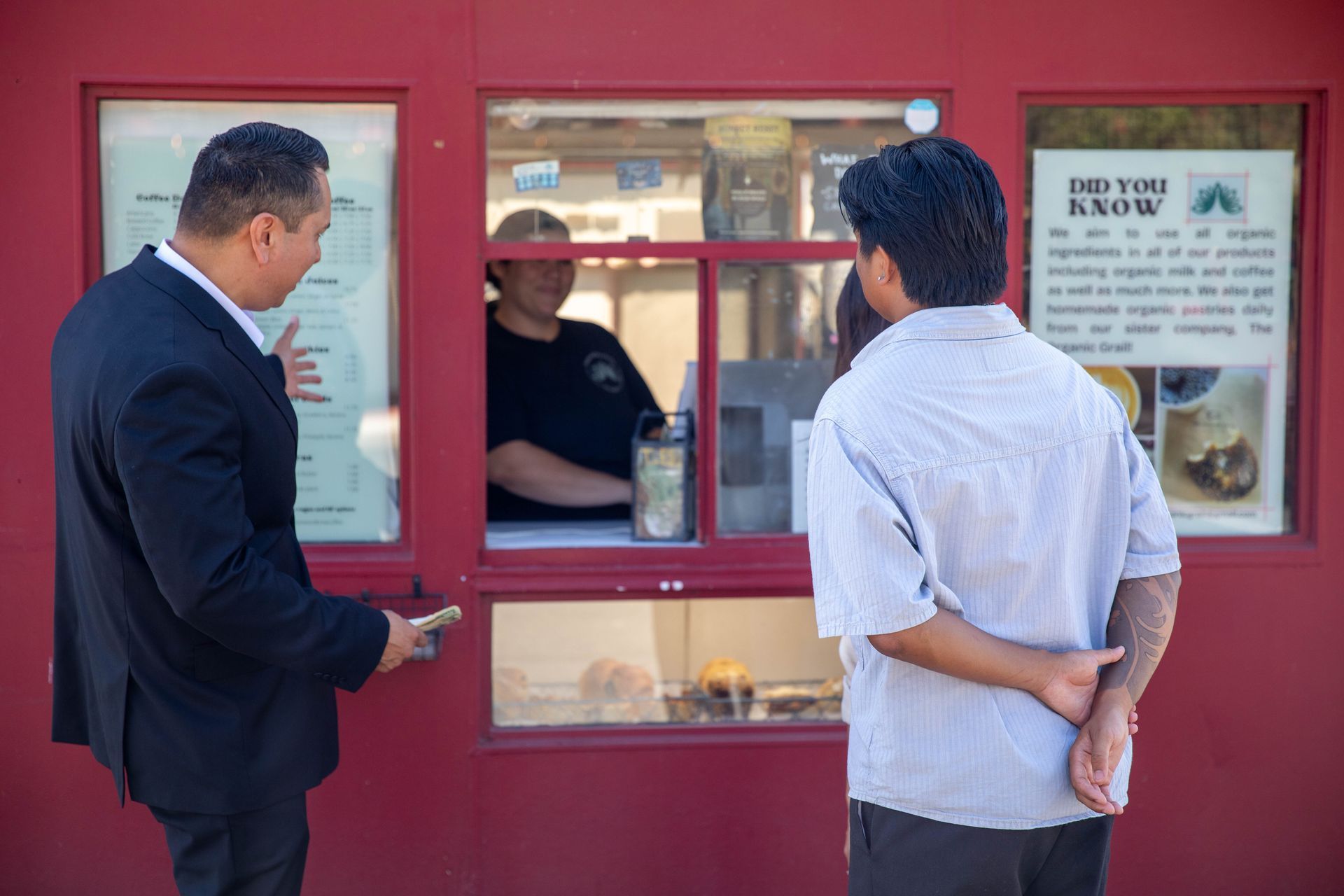 Two men at a red service window speaking with an attendant inside an office.