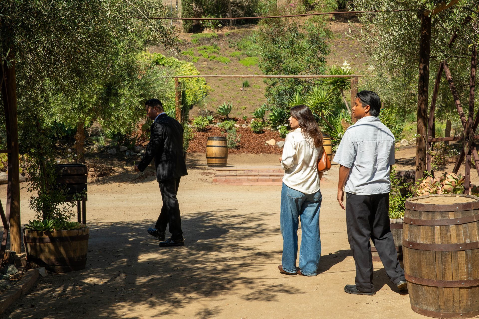 Three people standing on a sunlit garden path with trees and potted plants around them