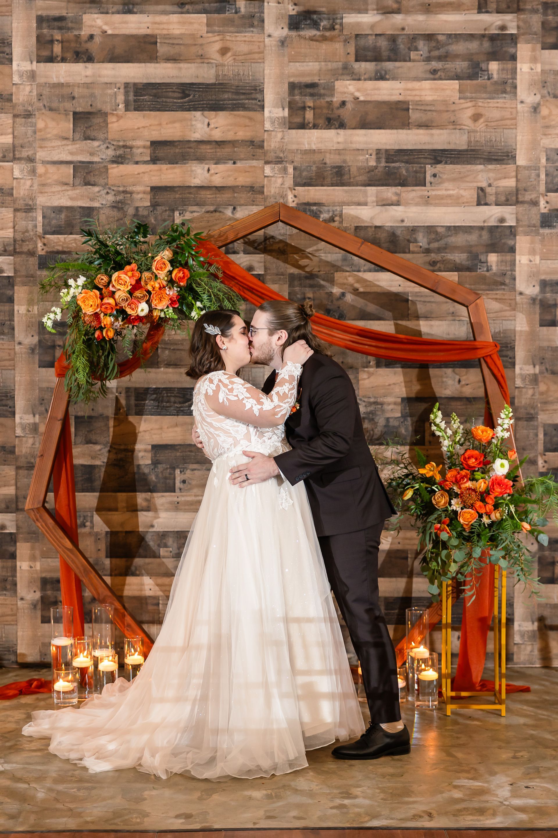 Bride and groom kissing under a floral hexagon arch at a rustic indoor wedding venue
