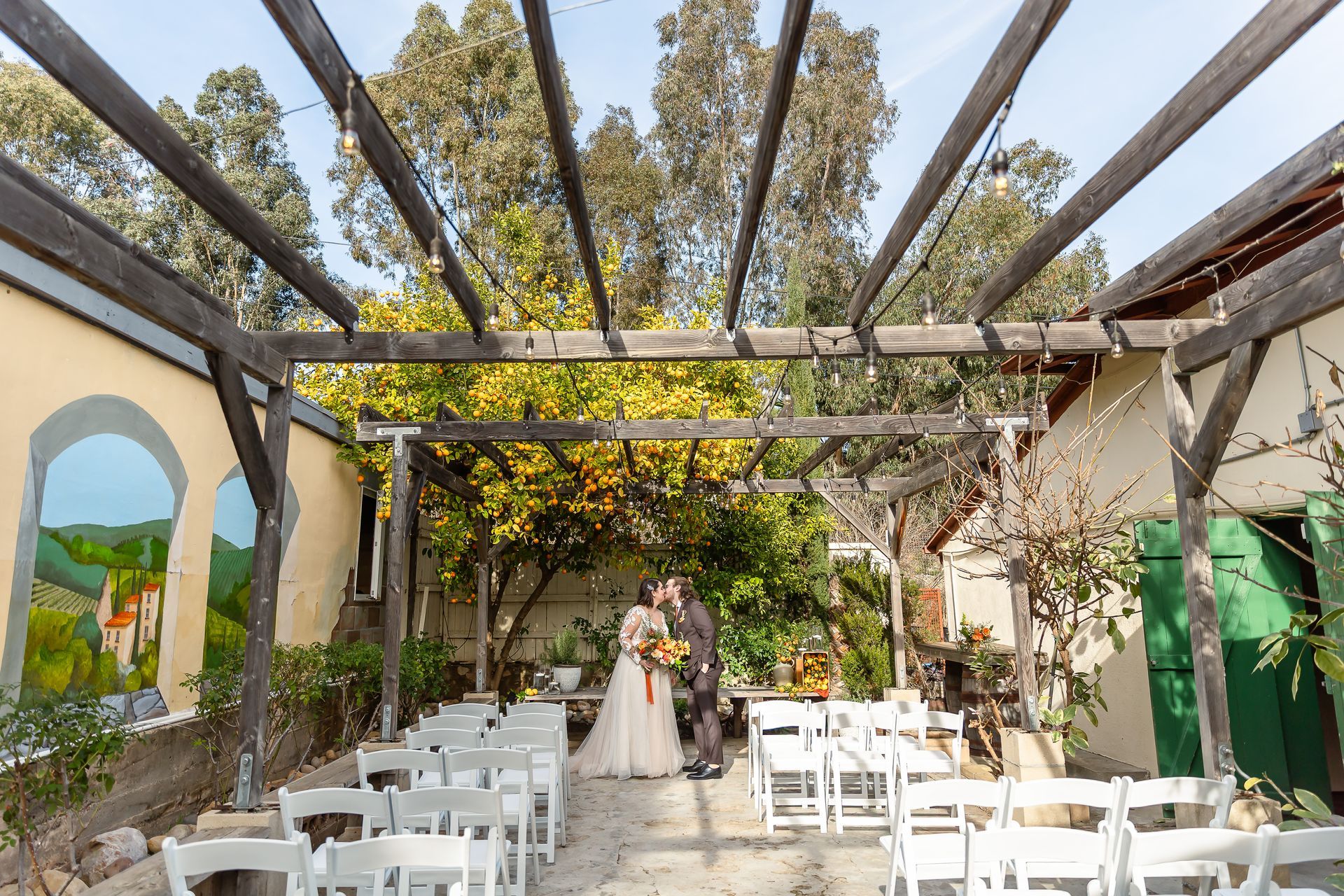 Open-air wedding ceremony under a vine-covered pergola, with white chairs and a couple at the altar